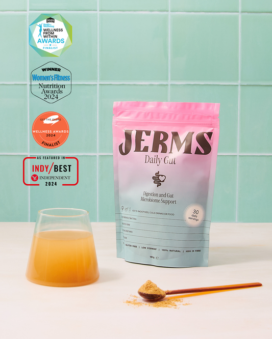 Jerms Daily Gut supplement package with a glass of juice and spoonful on a light surface against a green tiled wall.